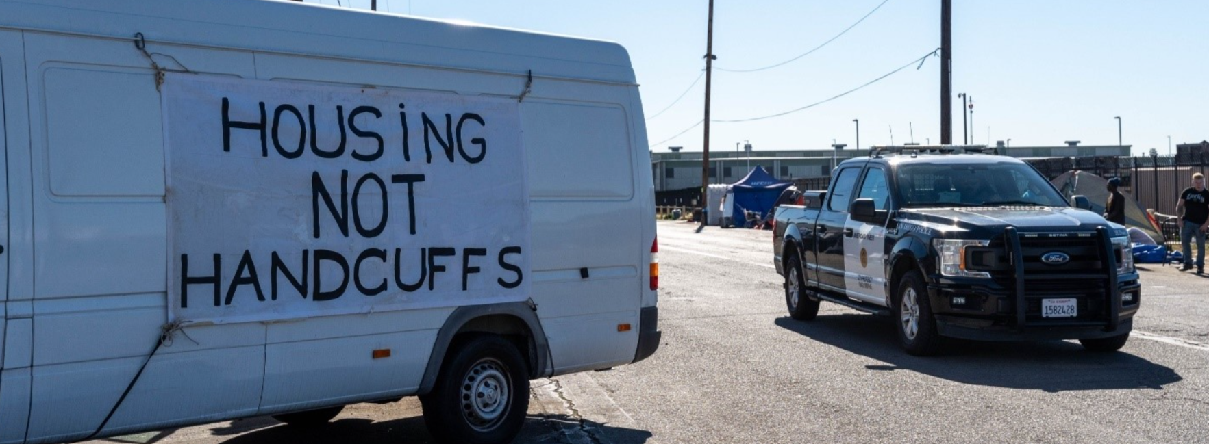 Photo of a van with a banner reading Housing Not Handcuffs, Police vehicle in background